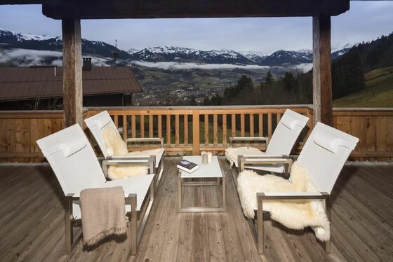Balkon vor Wohnzimmer Holzbalkon mit vier weißen Sesseln und Blick auf schneebedeckte Berge und ein Tal.