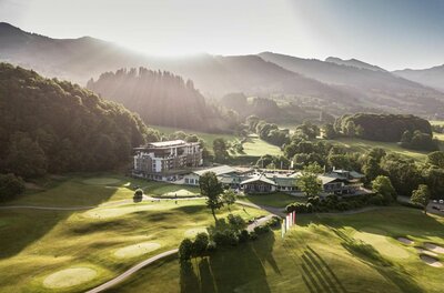 Luftaufnahme eines Hotels mit Golfplatz vor bewaldeten Hügeln bei Gegenlicht.