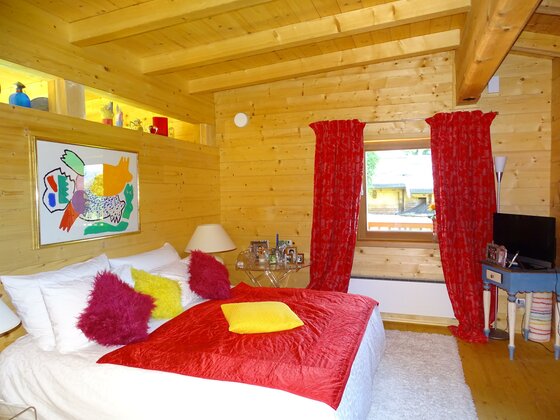 Schlafzimmer in einer Holzhütte mit Doppelbett, roten Vorhängen und einem bunten Wandbild.