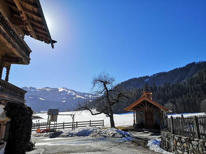 Hennleiten im Winter Alpines Holzhaus und kleine Kapelle vor verschneiter Berglandschaft unter blauem Himmel.
