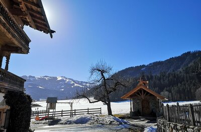 Alpines Holzhaus und kleine Kapelle vor verschneiter Berglandschaft unter blauem Himmel.