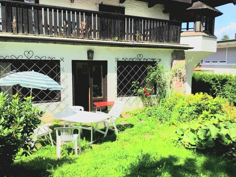 Garten mit weißen Kunststoffmöbeln, Sonnenschirm und einer Terrasse vor einem Haus mit Holzbalkon.