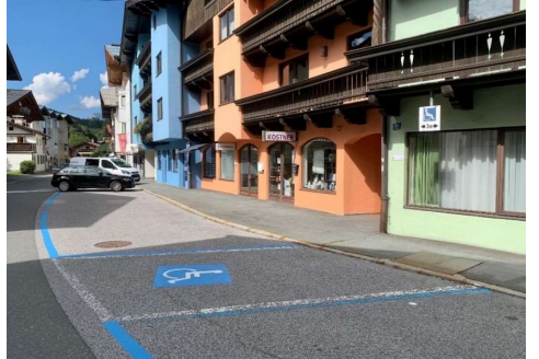 Eine ruhige Straße mit bunten Gebäuden und einem klaren Himmel. Parkplätze sind deutlich markiert, einschließlich eines Behindertenparkplatzes.