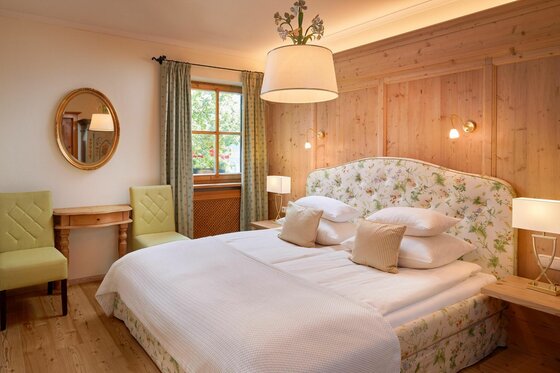 Doppelbett mit geblümtem Kopfteil in einem Zimmer mit Holzvertäfelung, Spiegel und zwei grünen Stühlen.