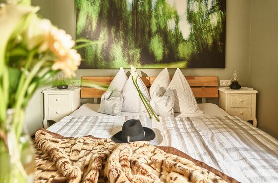 Schlafzimmer im Kitzhome City Apartment Doppelbett mit hellen Kissen, Hut auf der Decke und großem Waldbild an der Wand.