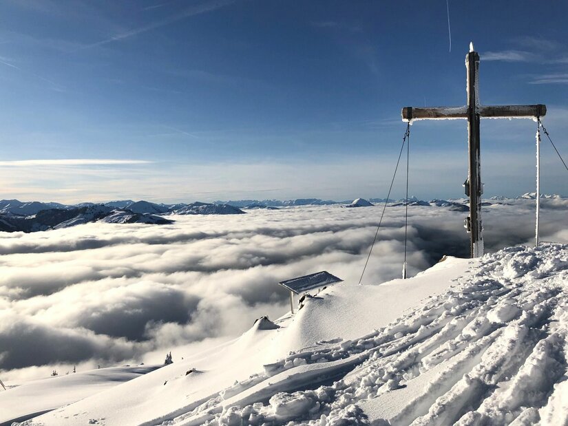 Gipfelkreuz auf dem verschneiten Hochetzkogel über einer geschlossenen Wolkendecke unter blauem Himmel.