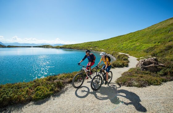 Zwei Mountainbiker fahren auf einem Schotterweg an einem blauen Bergsee unter klarem Himmel.