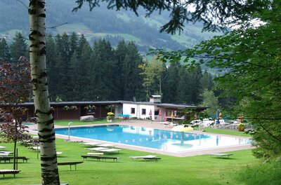 Freibad mit blauem Becken, Liegewiese und Holzgebäuden vor bewaldetem Berghang.