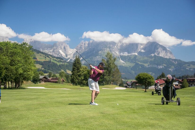 Ein Mann beim Golfschwung auf einem grünen Golfplatz vor einer alpinen Bergkulisse.