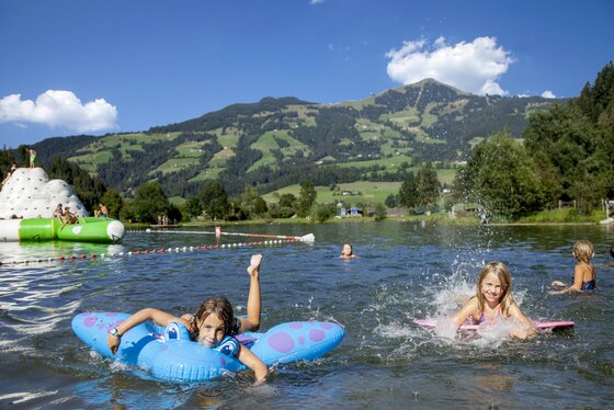 Kinder schwimmen in einem See mit bunten Schwimmringen. Im Hintergrund sieht man grüne Hügel und blauen Himmel.