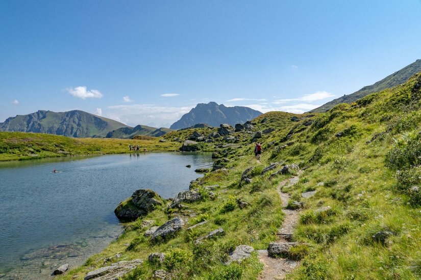 Wanderweg am Torsee Ein schmaler Wanderpfad führt an einem Bergsee vorbei durch eine grüne Alpenlandschaft mit Berggipfeln im Hintergrund.
