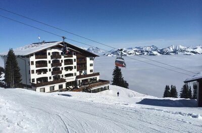 Ein mehrstöckiges Berghotel neben einer Skipiste und einem Sessellift über einer Wolkendecke im Winter.