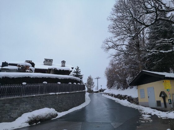 Verschneite Straße am Bauernhaus-Museumsweg mit Holzhäusern und Seilbahnmast im Hintergrund.