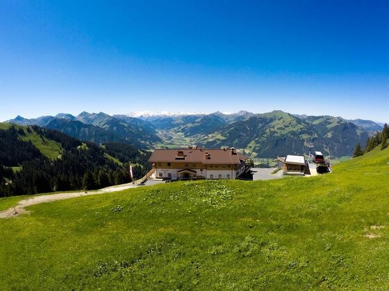 Alpengasthof auf einer grünen Bergwiese vor einem weiten Gebirgspanorama unter blauem Himmel.