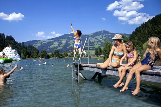 Ein Schwimmbad mit vielen Kindern und Familien am Wasser. Im Hintergrund sind grüne Berge und ein blauer Himmel zu sehen.