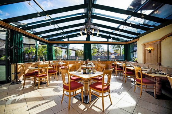 Ein helles Restaurant mit großen Fenstern und einem Glasdach. Die Tische sind sauber gedeckt und es gibt Holzstühle mit roten Polstern.