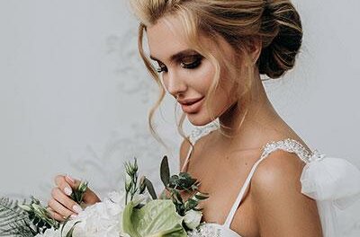 Eine Braut mit elegantem Haar und einem wunderschönen Blumenstrauß steht vor einem neutralen Hintergrund. Ihr Kleid hat zarte Details und strahlt eine romantische Atmosphäre aus.
