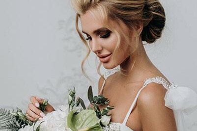 Galery Eine Braut mit elegantem Haar und einem wunderschönen Blumenstrauß steht vor einem neutralen Hintergrund. Ihr Kleid hat zarte Details und strahlt eine romantische Atmosphäre aus.