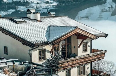 Alpines Wohnhaus mit Holzbalkon und schneebedecktem Dach vor verschneiter Bergkulisse.