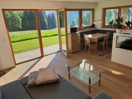 Wohnzimmer mit Sofa, Glastisch und Essecke vor großen Glasfronten mit Blick auf eine grüne Wiese.