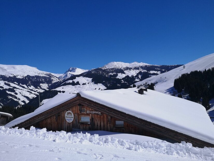 Schneebedeckte Holzhütte vor verschneiten Berggipfeln unter blauem Himmel.