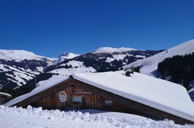 Bruggeralm im Winter Schneebedeckte Holzhütte vor verschneiten Berggipfeln unter blauem Himmel.