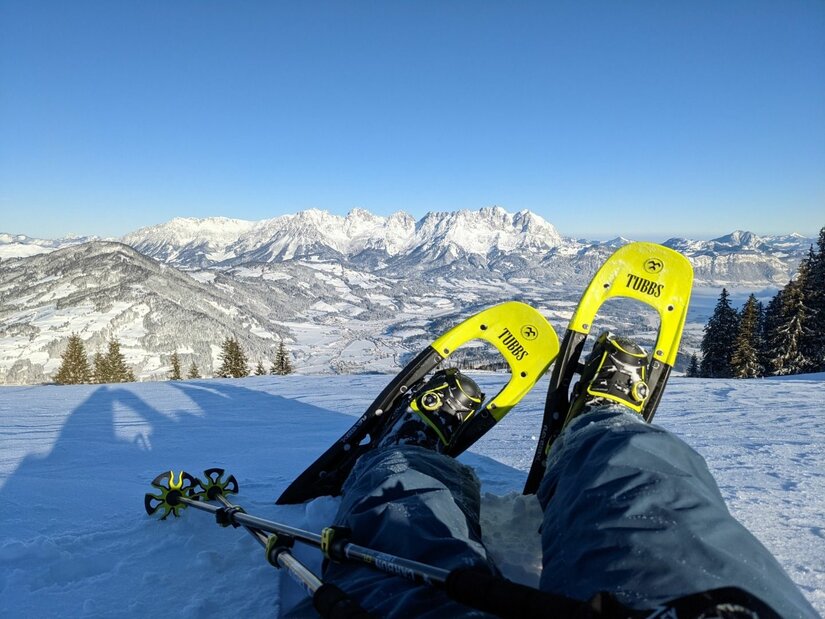 Schneeschuhwandern mit Bergblick Person mit gelben Schneeschuhen sitzt im Schnee vor einem verschneiten Bergpanorama bei blauem Himmel.