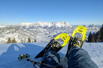 Schneeschuhwandern mit Bergblick Person mit gelben Schneeschuhen sitzt im Schnee vor einem verschneiten Bergpanorama bei blauem Himmel.