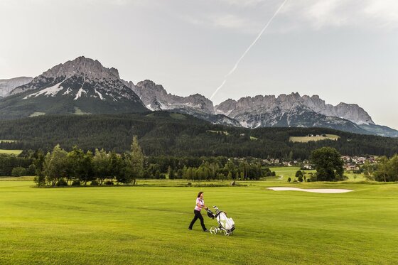 Ellmau_Wilder Kaiser Panorama_Wilder Kaiser Eine Person geht mit einem Golfwagen über ein grünes Golfplatz. Im Hintergrund sind beeindruckende Berge zu sehen.