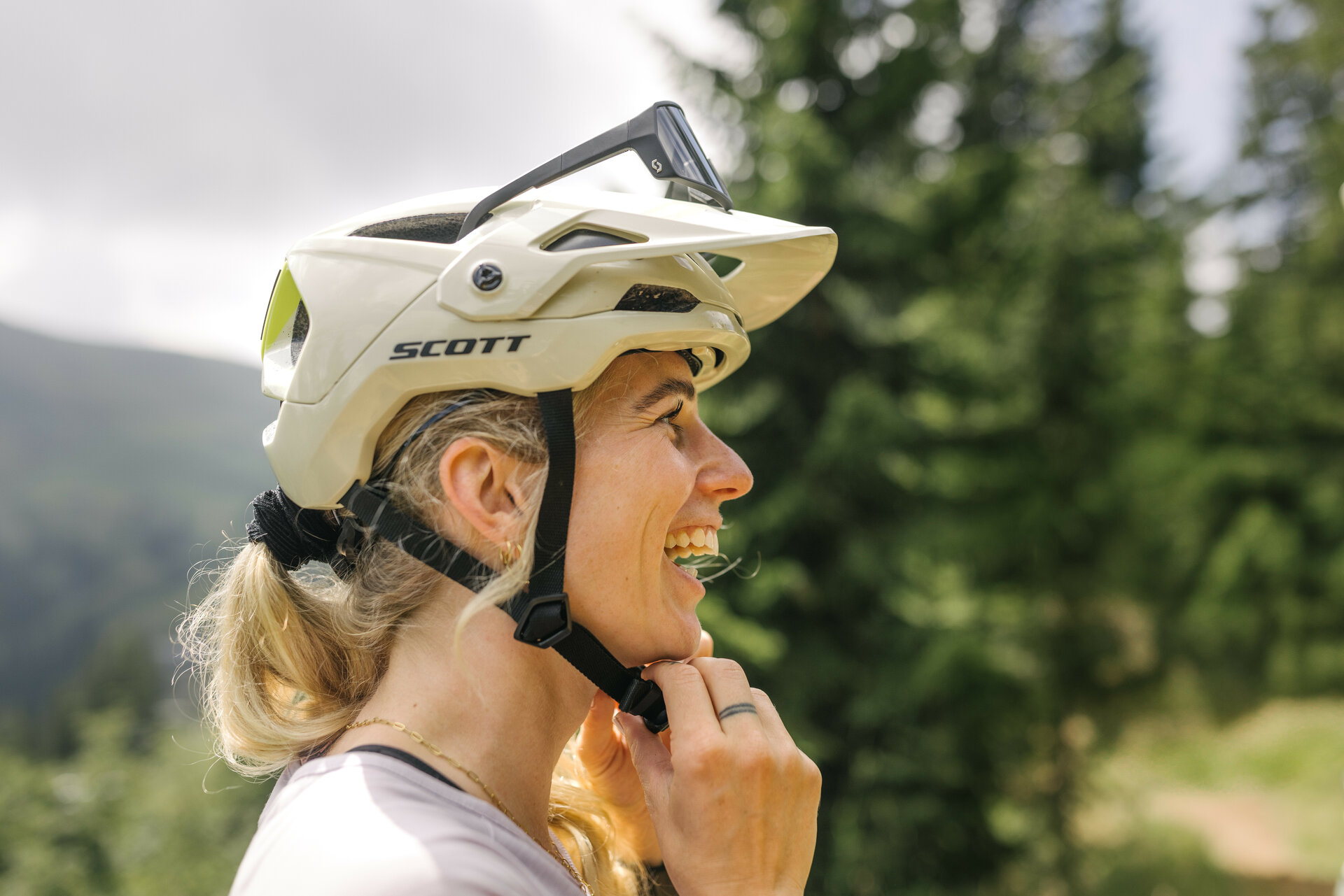360 Flow Trails in Kitzbühel: a smiling woman wearing a bike helmet stands amid green trees under a cloudy sky.