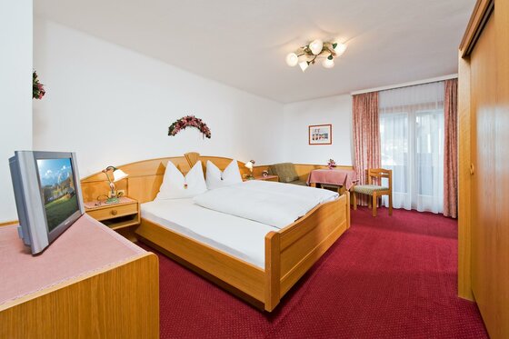 Doppelzimmer mit Holzmöbeln, rotem Teppichboden, Fernseher und Sitzecke vor einem Fenster.