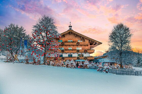 Rasmushof_Winter_2022_23-online Ein schneebedecktes Haus in den Bergen, umgeben von winterlichen Bäumen. Der Himmel leuchtet in sanften Farben bei Sonnenuntergang.