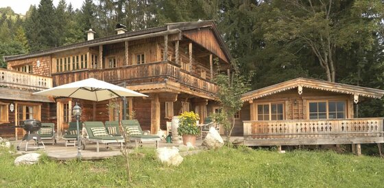 Zweistöckige Holzhütte mit Terrasse, Sonnenliegen, Sonnenschirm und Grill vor einem Waldrand.