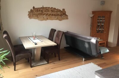 Ein Esstisch mit vier braunen Stühlen neben einem grauen Sofa in einem hellen Wohnraum.