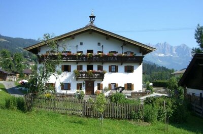 Ein traditionelles alpenländisches Haus mit Balkon und vielen Blumen. Im Hintergrund sind die Berge und ein blauer Himmel sichtbar.