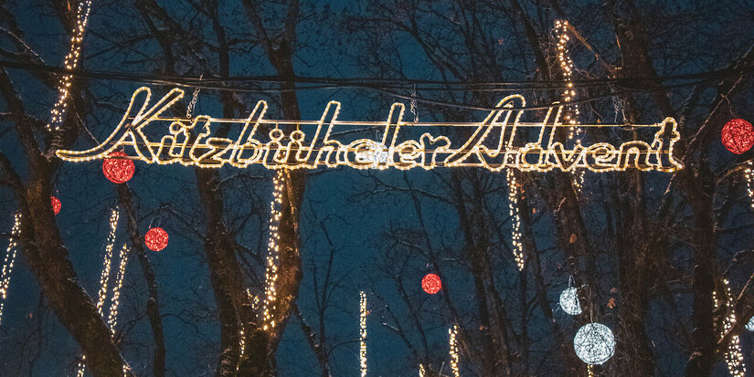 Die festlich beleuchtete Straße zeigt den Schriftzug 'Kitzbüheler Advent' zwischen glitzernden Lichtern und bunten Kugeln.