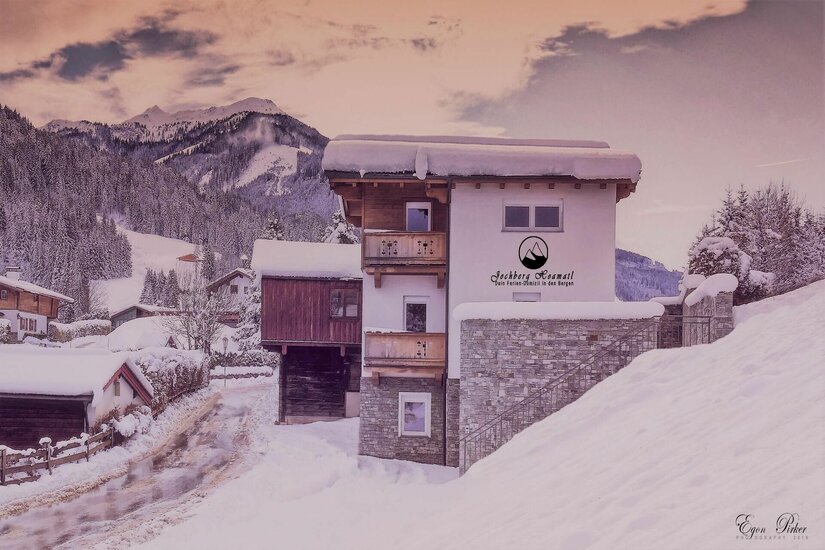 Schneebedecktes mehrstöckiges Ferienhaus mit Holzbalkonen vor bewaldeten Bergen im Winter.