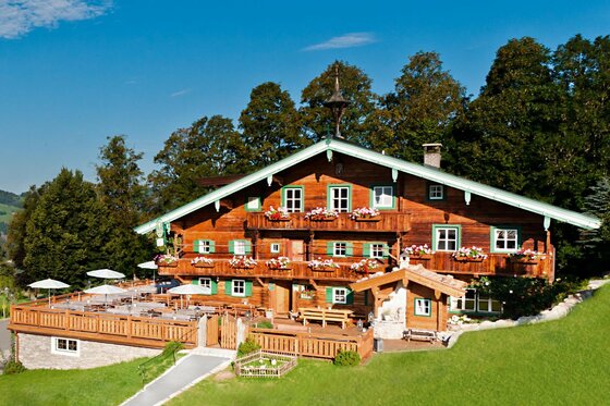Berggasthof Hagstein in Kitzbühel Alpiner Berggasthof aus dunklem Holz mit blumengeschmückten Balkonen und einer großen Sonnenterrasse vor Waldkulisse.