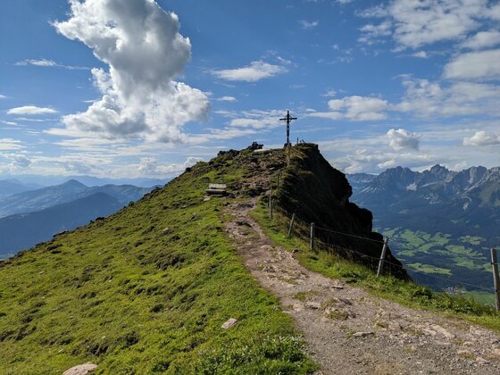 Ein schmaler Wanderweg führt über einen grasbewachsenen Bergrücken zu einem Gipfelkreuz unter blauem Himmel.