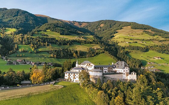 Luftaufnahme von Schloss Mittersill vor bewaldeten Berghängen und grünen Wiesen.