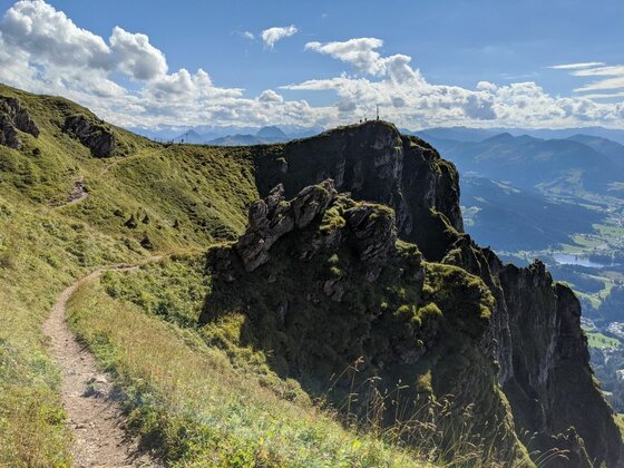 Schmaler Wanderpfad an einem grasbewachsenen Berghang mit Blick auf schroffe Felsformationen und ein Gipfelkreuz.