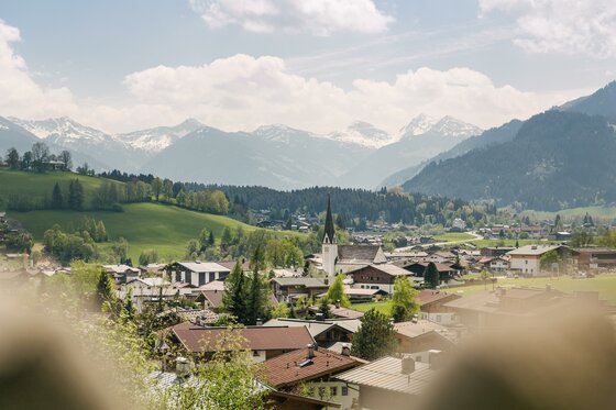 Panorama Reith im Frühling Blick über ein Alpendorf mit Kirchturm auf schneebedeckte Berggipfel unter bewölktem Himmel.