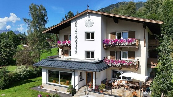 Außenansicht Hotel Garni Entstrasser Dreistöckiges Hotelgebäude mit weißen Wänden, Holzbalkonen mit Blumenschmuck und einer Terrasse im Erdgeschoss.