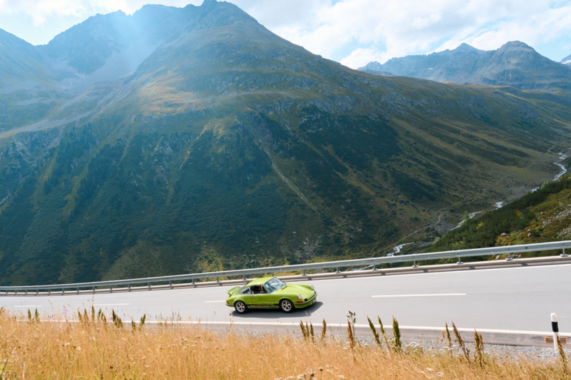 Ein grüner Sportwagen fährt auf einer asphaltierten Passstraße vor einer alpinen Bergkulisse.