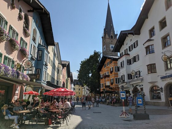 Belebte Fußgängerzone mit farbenfrohen Häuserfassaden und einem spitzen Kirchturm im Hintergrund.