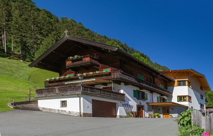 Alpiner Ferienhof mit dunkler Holzfassade, Balkonen und weißem Erdgeschoss vor bewaldetem Hang.