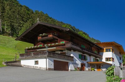 Alpiner Ferienhof mit dunkler Holzfassade, Balkonen und weißem Erdgeschoss vor bewaldetem Hang.