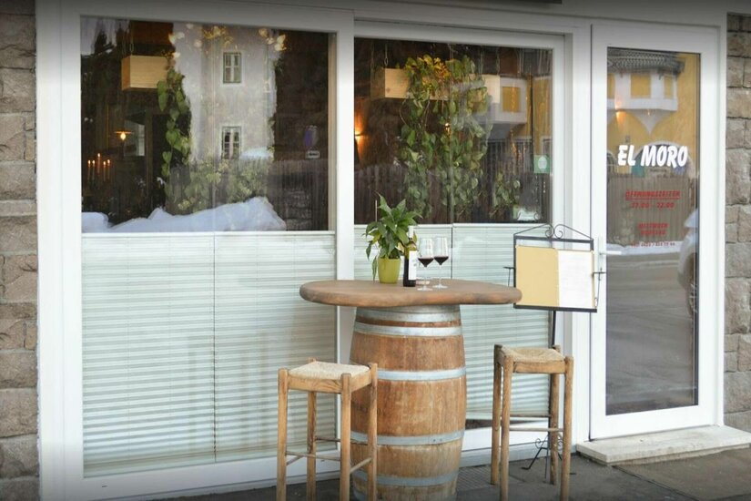 Außenbereich Pizzeria El Moro Außenansicht eines Restaurants mit einem Weinfass als Stehtisch, zwei Barhockern und Weingläsern vor einem Fenster.