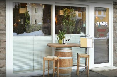 Außenansicht eines Restaurants mit einem Weinfass als Stehtisch, zwei Barhockern und Weingläsern vor einem Fenster.