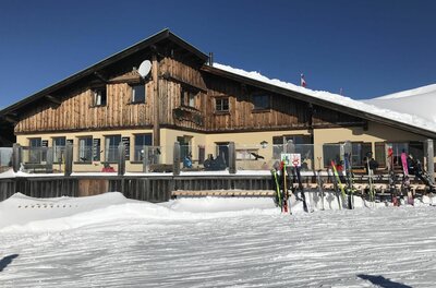Berghütte aus Holz und Stein im Schnee mit Skiern im Vordergrund unter blauem Himmel.